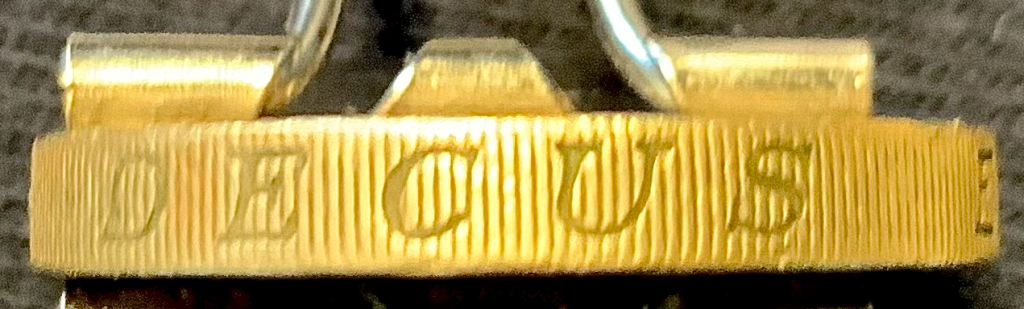 The milled edge of a British pound coin showing the first word of the term "decus et tutamen" with background grooved pattern.